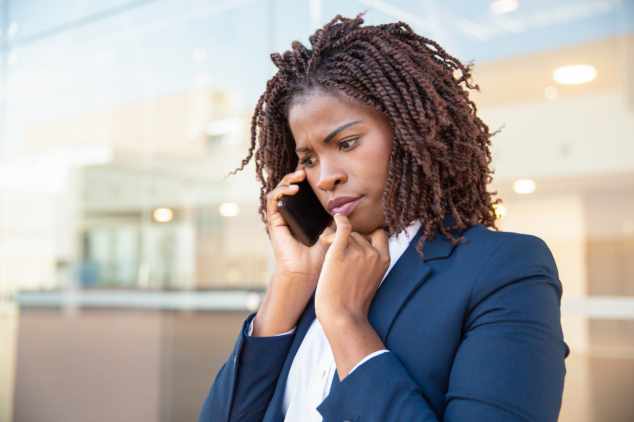 Frowning concerned manager speaking on cellphone outside. Young African American business woman standing near outdoor glass wall. Mobile phone talk concept